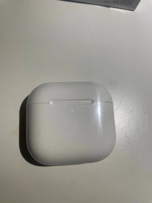 Aipods - Snygga trådlösa hörlurar i vit plast med matchande laddningsetui. Hörlurarna har en stilren och modern design med rundade former och diskreta detaljer. Perfekta för musik, poddar och samtal på språng.