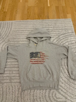 Grå hoodie från Denim & Supply - Snygg grå hoodie från Denim & Supply Ralph Lauren med amerikansk flagga i sliten look på bröstet. Klassisk känguruficka och huva med dragsko. Perfekt för dig som gillar streetwear och vill ha en avslappnad stil.