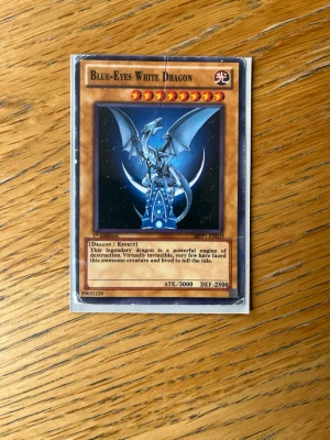 Yu-Gi-Oh! Blue-Eyes White Dragon - Säljer ett samlarkort från Yu-Gi-Oh! med motivet Blue-Eyes White Dragon. Kortet har en blå och vit drake mot en mörk bakgrund och är tryckt på klassiskt spelkortsmaterial. Det är en 1st Edition med ATK 3000 och DEF 2500.