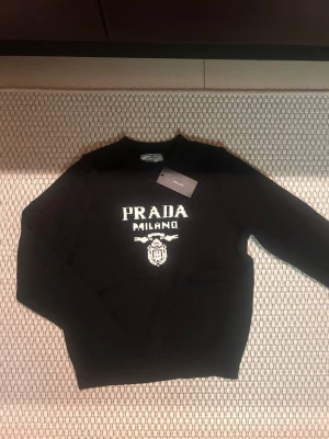 Prada sweater - Svart stickad tröja från Prada med vit logotyp och texten 'PRADA MILANO' framtill. Tröjan har rund halsringning och långa ärmar. Klassisk och stilren design med ribbstickade muddar vid ärmslut och nederkant.