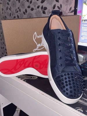 Mörkblå sneakers med nitar - Säljer ett par mörkblå sneakers från Christian Louboutin i mocka med svarta nitar på tån och den klassiska röda sulan. Skorna har vita kanter på sulan och matchande mörkblå skosnören. Perfekta för dig som vill sticka ut med en edgy och lyxig stil.