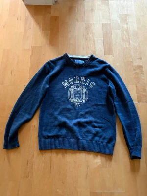 Marinblå tröja från Morris - Snygg marinblå tröja från Morris med vit logga och emblem på bröstet. Tröjan har rund halsringning, långa ärmar och ribbade muddar vid ärmslut och nederkant. Perfekt för en stilren och avslappnad look. Storlek medium