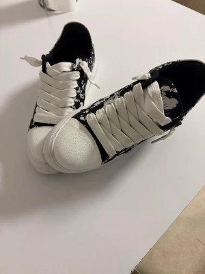 Svarta och vita sneakers med mönster - Säljer ett par snygga sneakers i svart och vitt med ett unikt stänkmönster. Skorna har breda vita snören, vit tå och sula samt låg profil. Perfekta för dig som vill sticka ut med din stil.