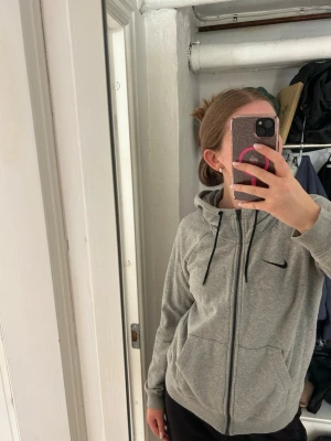 Grå zip hoodie från Nike - Säljer en grå zip hoodie från Nike med svart logga på bröstet. Hoodien har dragkedja framtill, två fickor och justerbar huva med svarta snören. Perfekt för chill dagar eller träning. Klassisk och enkel design som passar till det mesta.