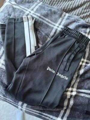 Palm Angels byxor - Snygga svarta mjukisbyxor från Palm Angels med vita ränder längs sidorna och logga tryckt på benet. Byxorna har resår i midjan och dragsko samt fickor på sidorna. Perfekta för en chill och trendig look. Palm angels lappen på insidan är lös på en sida enligt bild