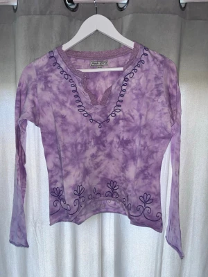 Lila långärmad topp från Fresh Spirit - Lila långärmad topp från Fresh Spirit med batikmönster och broderade detaljer längs v-ringningen och nederkanten. Toppen har spetskant vid halsen och är tillverkad i ett mjukt material som känns skönt mot huden. Perfekt för dig som gillar unika plagg!