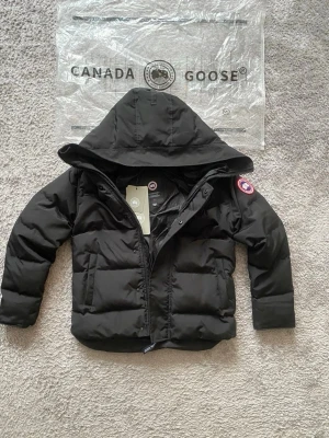 Canada Goose - MacMillan Parka - Svart dunjacka från Canada Goose med huva och klassisk logotyp på ärmen. Jackan har dragkedja framtill och två stora fickor. Perfekt för kalla dagar och riktigt snygg streetstil.