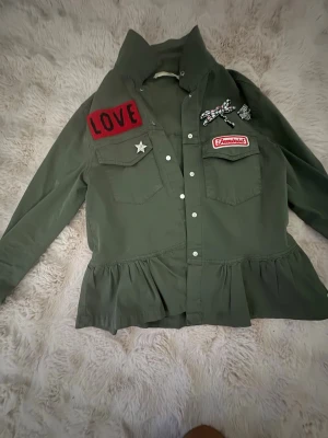 Grön skjorta med volang och patches - Stilren grön skjorta med volang nedtill och flera coola patches, bland annat en röd 'LOVE'-patch, en 'Feminist'-patch och dekorerade broscher. Skjortan har långa ärmar, knappar framtill och två bröstfickor. Perfekt för dig som gillar statement-plagg.