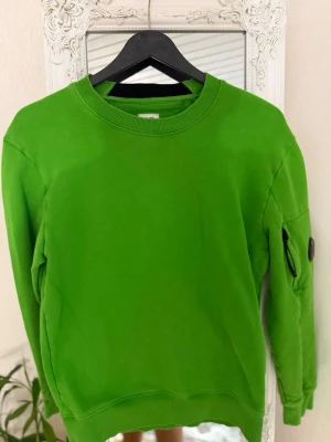 Grön sweatshirt från C.P. Company - Snygg grön sweatshirt från C.P. Company med rund halsringning och ribbade muddar. Tröjan har en ikonisk svart linsdetalj på ärmen och en liten ficka med dragkedja. Perfekt för dig som gillar streetwear och vill sticka ut. Priset kan sänkas 