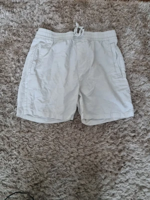 Vita shorts med snörning - Säljer ett par vita shorts med elastisk midja och snörning framtill. Shortsen har två sidofickor och är tillverkade i ett mjukt bomullstyg, perfekta för varma dagar. Passformen är normal och de har en enkel, clean look.