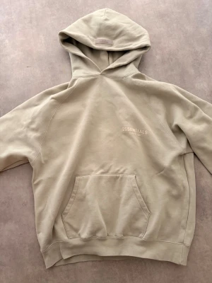 Sage green Essentials Fear of God hoodie - Snygg sage green hoodie från Essentials Fear of God med broderad logga på bröstet. Hoodien har en stor huva, känguruficka och är gjord i mjukt bomullsmaterial. Perfekt för en avslappnad och trendig stil.