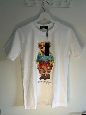 Vit Polo Bear t-shirt från Ralph Lauren - Säljer en vit t-shirt från Ralph Lauren med den ikoniska Polo Bear-trycket framtill. Björnen har solglasögon, grön jacka, röd tröja knuten runt midjan och bruna byxor. T-shirten har rund hals och korta ärmar. Perfekt för dig som gillar streetstyle och klassiska prints.