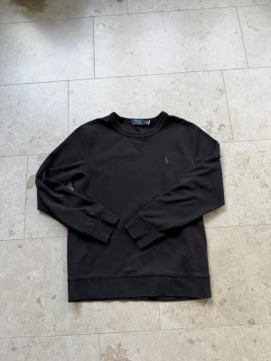 Svart sweatshirt från Polo Ralph Lauren - Klassisk svart sweatshirt från Polo Ralph Lauren med rund halsringning och blå broderad logga på bröstet. Tröjan har ribbade muddar vid ärmslut och nederkant samt en dekorativ söm vid halsen. Perfekt för en avslappnad och stilren look.