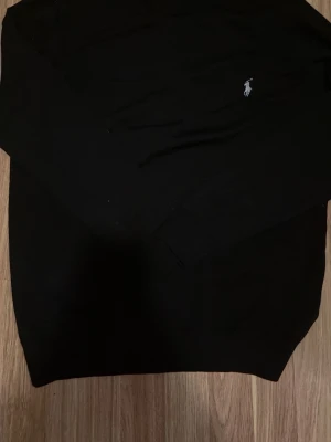 Svart stickad tröja från Polo Ralph Lauren - Klassisk svart stickad tröja från Polo Ralph Lauren med den ikoniska broderade loggan på bröstet. Tröjan har rund halsringning och långa ärmar, perfekt för lager på lager och en clean look.