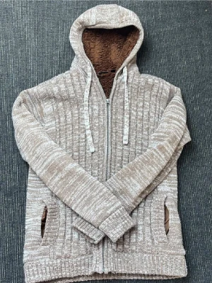 Beige stickad hoodie med dragkedja - Mysig beige stickad hoodie med dragkedja och huva. Utsidan har ett melerat mönster i beige och vitt, insidan av huvan är fodrad med mjukt brunt tyg. Två fickor framtill och ribbade muddar vid ärmslut och nederkant. Perfekt för chill dagar.