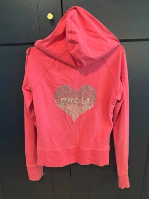 Rosa hoodie med strassdetaljer - Snygg rosa hoodie från Guess med dragkedja framtill och huva. På ryggen finns ett hjärta i strass med Guess-loggan och mindre strassdetalj på bröstet. Hoodien har långa ärmar och ribbade muddar. Perfekt för en trendig och avslappnad stil.