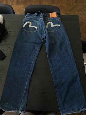 Blå raka jeans med broderade fickor - Säljer ett par blå jeans med raka ben och broderade detaljer i vitt på bakfickorna. Jeansen har klassisk femficksdesign och kontrastsömmar. Materialet är kraftig denim som ger en tidlös look.