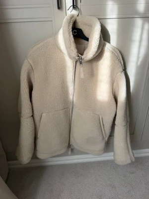 Beige/vit teddyjacka med hög krage - Mysig beige teddyjacka med dragkedja framtill och två stora fickor. Jackan har en hög krage och är gjord i fluffigt, mjukt material som håller dig varm. Perfekt för kyliga dagar och ger en avslappnad, trendig look.