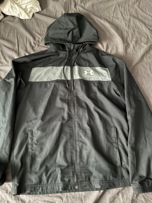 Svart vindjacka från Under Armour - Snygg svart vindjacka från Under Armour med grått parti över bröstet och logga. Jackan har huva med dragsko, hel dragkedja framtill och två sidofickor. Tillverkad i lätt och tåligt material, perfekt för blåsiga dagar.