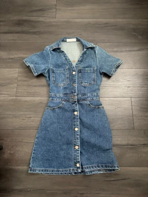 Kort jeansklänning med knappar - Säljer en snygg kort jeansklänning med knappar framtill, två bröstfickor och korta ärmar. Klänningen har klassisk skjortkrage och markerad midja för en cool och avslappnad look. Perfekt till sneakers eller boots!