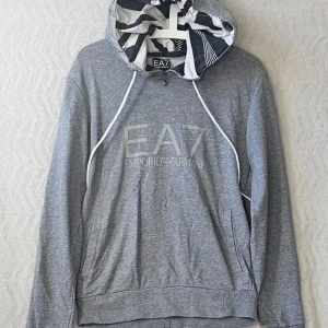 Grå hoodie från EA7 Emporio Armani - Snygg grå hoodie från EA7 Emporio Armani med vit logga på bröstet. Huvan har ett coolt svartvitt mönster på insidan och vita dragsnören. Hoodien har dragkedja upptill och två fickor framtill. Perfekt för en avslappnad och trendig look.
