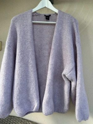 Ljuslila stickad kofta - Mysig och oversized stickad kofta i ljuslila färg. Koftan har öppen front utan knappar och långa ärmar med nedhasad axelsöm. Tillverkad i ett mjukt, fluffigt material som ger en avslappnad look.
