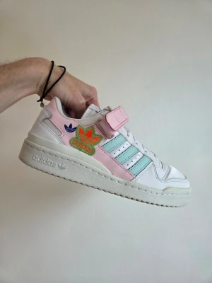 Adidas Forum Low pastell sneakers - Snygga Adidas Forum Low sneakers i vitt läder med pastellrosa och ljusblå detaljer. Skorna har kardborreband över vristen, klassiska tre ränder på sidan och färgglad Adidas-logga. Perfekta för dig som gillar retrovibbar och streetstyle.
