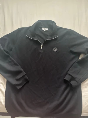 Svart half-zip hoodie Moncler - Säljer en svart hoodie från Moncler med half-zip dragkedja och hög krage. Tröjan har ett Moncler-märke på bröstet och är tillverkad i mjukt material. Långa ärmar och ribbade muddar ger en clean look. Perfekt för dig som gillar stilrena plagg. OBS: storelk S och inte L