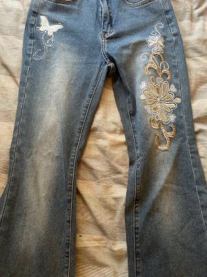 Blå utsvängda jeans med fjärilsbrodyr - Säljer ett par blå jeans med utsvängda ben och snygg broderad fjäril med pärldetaljer.  . Perfekta för dig som gillar unikt.