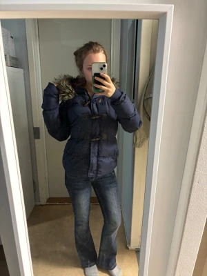 Demin co kacka - Snygg mörkblå pufferjacka med huva och fuskpälsdetalj. Jackan har träknappar och dragsko i midjan för en schysst passform. Insidan av huvan är fodrad i rött tyg. Perfekt för kalla dagar och riktigt mysig att bära.