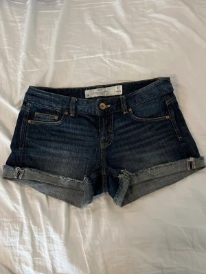 Mörkblå jeansshorts med upprullad kant - Snygga mörkblå jeansshorts med upprullade benslut och slitna detaljer. Klassisk femficksmodell med knapp och dragkedja framtill. Tillverkade i denim med en skön passform, perfekta för sommardagar.