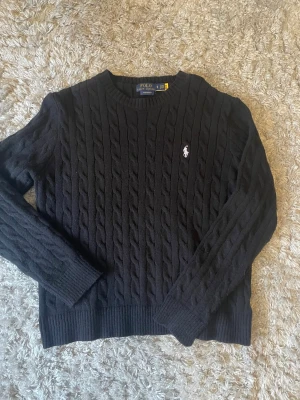 Polo - Klassisk svart kabelstickad tröja från Ralph Lauren. Mjukt material med ikonisk broderad häst på bröstet. "Custom Fit". Perfekt för alla tillfällen.