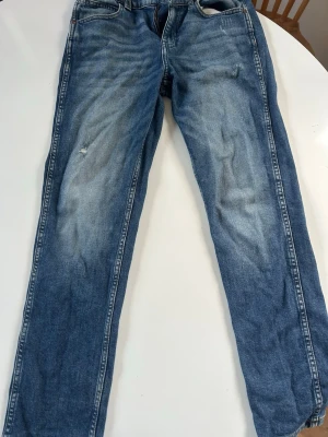 Blå raka jeans med slitningar - Säljer ett par klassiska blå jeans med raka ben och snygga slitningar på framsidan. Jeansen har fem fickor, kontrastsömmar och en cool vintage tvätt. Bakfickorna har dekorativa sömmar. Perfekta för en avslappnad och trendig look.