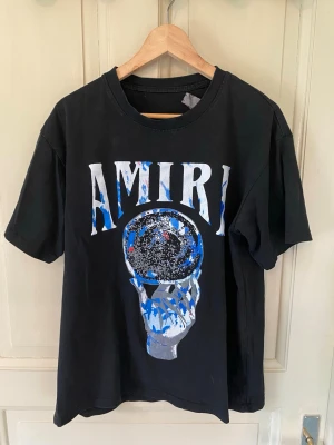 Svart AMIRI t-shirt  - Svart t-shirt från AMIRI med stort grafiskt tryck framtill i blått, vitt och silver. Klassisk rund hals och korta ärmar. Materialet ser ut att vara mjuk bomull och passformen är normal. Perfekt för dig som gillar streetwear och coola prints. 