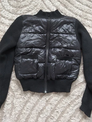 Svart pufferjacka från Gina Tricot - Snygg svart pufferjacka från Gina Tricot med stickade ärmar och krage. Jackan har två stora fickor framtill och dragkedja hela vägen. Kroppen är i glansigt, vadderat material medan ärmar och krage är ribbstickade för extra komfort.