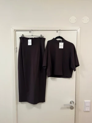 Brunt set med kjol och topp - Snyggt tvådelat set i mörkbrunt med en lång, rak kjol och en topp med vida, halvlånga ärmar och hög krage. Setet har minimalistisk design och är gjort i ett mjukt, stretchigt material som ger en skön känsla. Perfekt för dig som gillar stilrena plagg.