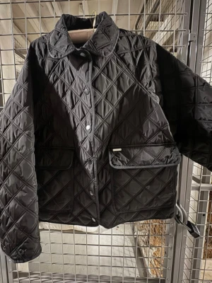 Svart quiltad jacka med krage - Från hemsidan: Quilted Femme Jacket har en vattenavvisande finish (Bionic-Finish® ECO) och en klassisk quiltad design med lyxig semi-shine. Quiltad Femme Jacket har ett boxigt snitt, knappstängning, justerbar dragsko och tryckknappar vid ärmslut. Jackan har smarta ”2-i-1-fickor” på varje sida, vilket innebär att ena fickan nås ovanifrån och den annan nås från sidan.  Material: Skal: 100 % polyester Foder: 100 % polyester Fyllning: 100 % återvunnen polyester  Använd men inga synliga tecken på anv