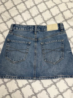 Kort jeanskjol med fickor - Säljer en klassisk blå jeanskjol i kort modell med fem fickor och bälteshällor. Kjolen har en rak passform och är tillverkad i slitstarkt denimtyg. Perfekt att matcha med en t-shirt eller hoodie för en avslappnad look.