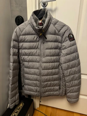 Ljusgrå dunjacka från Parajumpers - Snygg ljusgrå dunjacka från Parajumpers med quiltad design och hög krage. Jackan har dragkedja framtill, två sidofickor och en patch med logga på ärmen. OBS: Liten i storlek, kan absolut passa för M också.