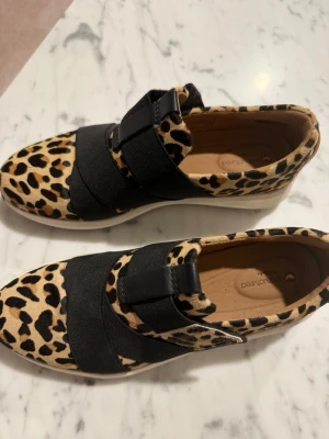 Leopardmönstrade sneakers med resårband - Snygga sneakers med leopardmönster i beige och svart. Skorna har breda svarta resårband över foten för en cool look och enkel påtagning. Yttermaterialet är textil med djurmönster och sulan är vit och tjock för extra komfort.