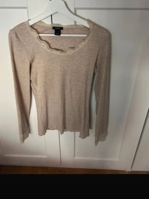 Beige långärmad topp med spets - Söt beige långärmad topp från Lindex med spetsdetaljer vid halsringningen och ärmsluten. Tillverkad i mjukt material och har en feminin, figurnära passform. Perfekt att styla med jeans eller kjol.
