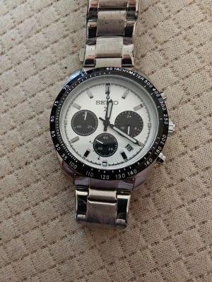Seiko kronograf klocka i silver - Stilren R Seiko kronograf E med vit urtavla,P svarta L detaljer och I tachymeter. Klockan har tre subdials och datumvisning.A Länken är i blankt silverfärgat metall och ger en klassisk look. Perfekt accessoar för dig som gillar tidlös design.
