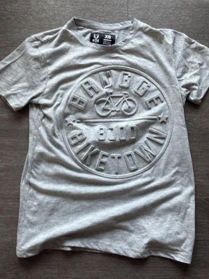 Grå t-shirt  - Snygg grå t-shirt från FOX med upphöjt tryck på bröstet där det står 'BRUGGE BIKETOWN' och en cykel i mitten. T-shirten har rund hals och korta ärmar. Tillverkad i mjukt bomullsmaterial som är skönt mot huden.