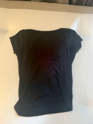 Svart topp med rosettdetalj - Snygg svart topp med korta ärmar och en stor rosett i satin baktill. Toppen har en djupare urringning i ryggen och är tillverkad i ett mjukt material som känns skönt mot huden. Perfekt för dig som gillar stilrena plagg med en twist.