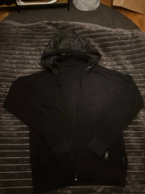 Indicode cardigan  - Svart cardigan från INDICODE med hel dragkedja framtill och huva i glansigt material. Hoodien har långa ärmar, ribbade muddar och fickor med dragkedja. Perfekt för en chill och stilren look. Slim fit 