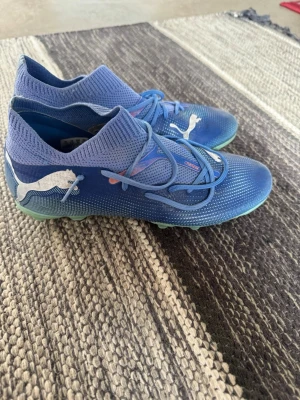 Blåa Puma fotbollsskor med snörning - Snygga blåa fotbollsskor från Puma med vit logga på sidan och stickad, strumpliknande ovandel. Skorna har snörning för bra passform och en lätt, flexibel sula med dobbar för spel på gräs. Perfekta för dig som vill ha stil och funktion på planen.