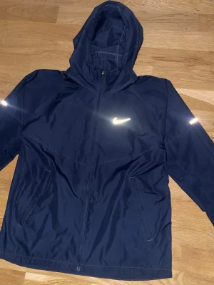 Nike Miler Windbreaker  - Snygg mörkblå vindjacka från Nike med reflekterande logga på bröstet och detaljer på ärmarna. Jackan har dragkedja framtill, två sidofickor och en praktisk huva. Tillverkad i lätt och tåligt material som står emot vind.