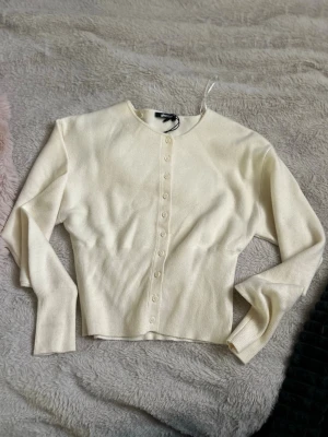 Krämvit ribbad kofta med knappar - Snygg krämvit kofta med rund hals och långa ärmar. Koftan har ribbade muddar och midja samt knappar hela vägen framtill. Perfekt att styla till jeans eller kjol för en clean look. (Ej använd) 