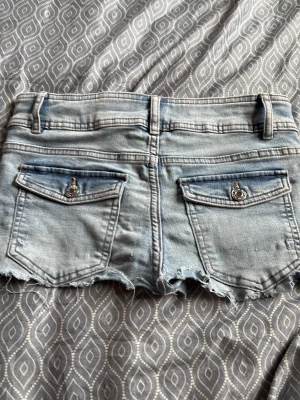 Ljusblå jeansshorts med rå kant - Snygga ljusblå jeansshorts med fransig, rå kant och klassiska fem fickor. Shortsen har dubbla knappar framtill och bakfickor med lock och knappdetalj. Perfekta för varma dagar och en avslappnad stil.
