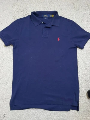 Marinblå pikétröja från Ralph Lauren - Klassisk marinblå pikétröja från Ralph Lauren med röd broderad logga på bröstet. Slim fit-modell med krage och knappar framtill. Perfekt till jeans eller shorts för en clean och stilren look. M men sitter som S